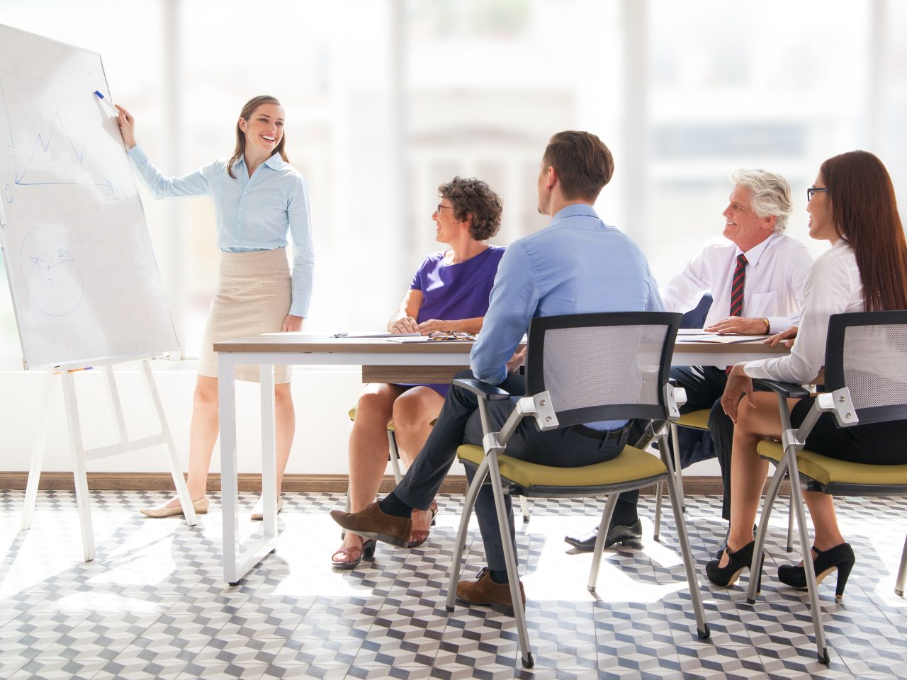 Cheerful young businesswoman pointing at whiteboard and explaining strategy. Confident business coach presenting project to staff. Colleagues listening to presenter. Business meeting concept